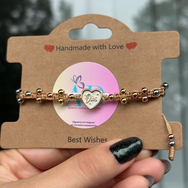 Pulsera tejida con perlas doradas, hilo café y un corazonistas que dice Dios