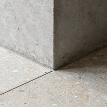 Macro architectural detail showing the clean intersection of a raw concrete column meeting a honed travertine floor tile, soft natural side-light.
