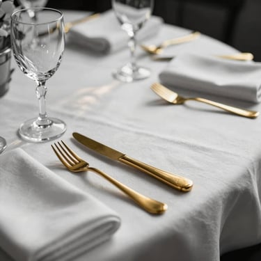 Close-up detail of a formally set place setting on a white linen tablecloth — gold-toned cutlery aligned precisely beside a folded napkin, crystal glassware catching afternoon window light, shallow depth of field, black-and-white tones with the warm gold of the cutlery as the single colour anchor