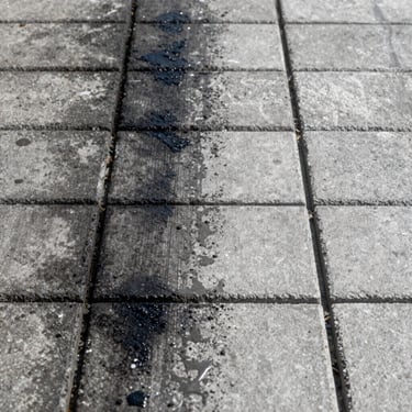 Split-frame close-up of concrete paving blocks: left half dark with black oil stains and embedded grime between joints, right half pressure-washed to clean light grey, sharp joint lines visible, bright daylight