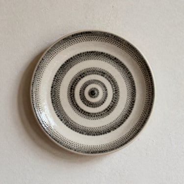 Decorative ceramic wall plate with black and white textured spiral dot painting, styled on a neutral plaster wall