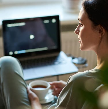 A woman relaxing at home watching a video on her laptop while holding a cup of hot tea.