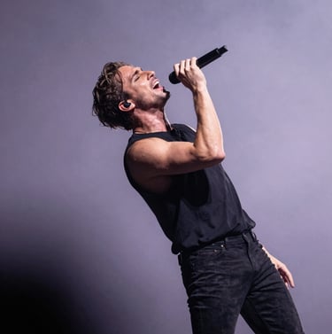 An action shot of Stefan Taylor mid-performance, leaning into a high note. The background is a soft lavender grey haze with dramatic dark charcoal shadows, capturing the artistic elegance of his movement.