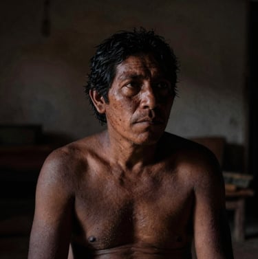 A contemplative, low-light photograph of a human figure in a South American / Argentine home, partially obscured by shadows. The composition focuses on the play of light on skin, using a palette of deep charcoal and muted earthy brown.
