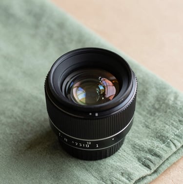An understated close-up of a vintage lens resting on a sage green cloth. The lighting is soft and professional, capturing the fine details of the glass against a muted sand surface.