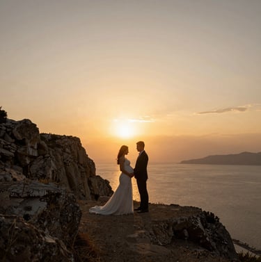 A wide angle outdoor shot of a bride and groom during a maternity shoot on a Bodrum cliffside at golden hour. The composition is minimal, focusing on the silhouette against the sunset. Warm mustard and charcoal tones in the landscape.