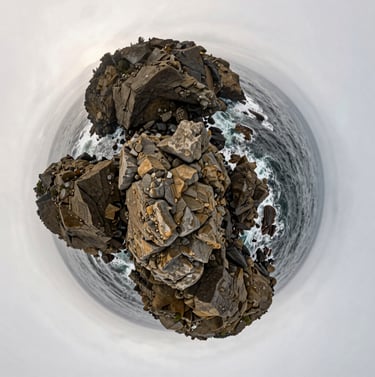 A distorted, 360-degree 'tiny planet' view of a rocky coastline in Oregon. The ocean is a deep muted blue-grey, and the rugged rocks have warm taupe textures under a soft off-white sky.