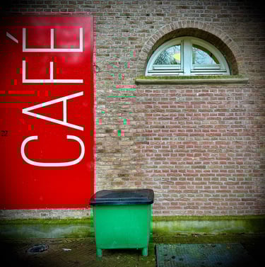 Vertical red cafe sign on a brick wall next to a green trash bin and arched window.