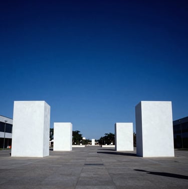 A wide-angle landscape photograph of a public plaza in North America featuring Bauhaus-style sculptures. The composition is grounded by large white rectangular blocks under a deep cerulean blue sky. Professional photography with high clarity and balance.