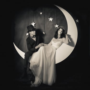 Vintage black and white wedding photo of a couple posing on a giant crescent moon prop with hanging stars.