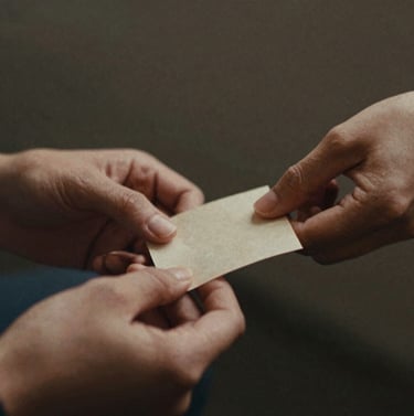 A soft-focus, intimate shot of two hands gently holding a small, worn piece of paper. The lighting is dim, suggesting a private, quiet moment. The aesthetic is film-like with visible grain. The colors are predominantly charcoal #3E352B and beige #D1B499.