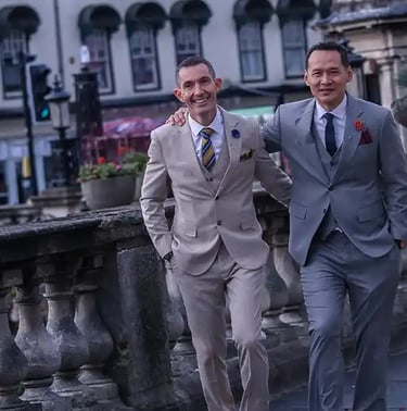 Two smiling men walking outdoors in stylish, tailored three-piece suits with ties and pocket squares.