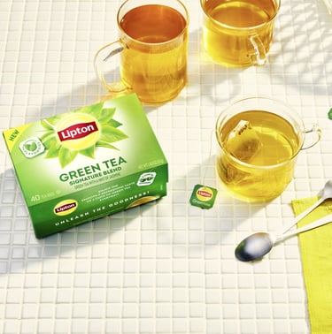 A box of Lipton Green Tea Signature Blend next to mugs of hot tea on a white tiled surface.
