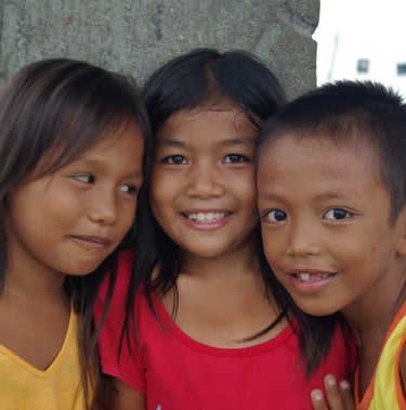 Three smiling Filipino children posing closely together outdoors in a rural village setting.