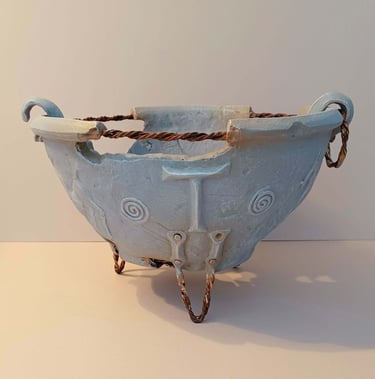 A fragmented, round bowl-like sculpture adorned with symbols and letters, finished in blue-grey hues