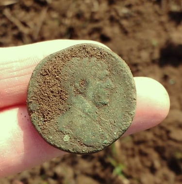 A large Roman coin with green patina, held out on two fingers of the finder.