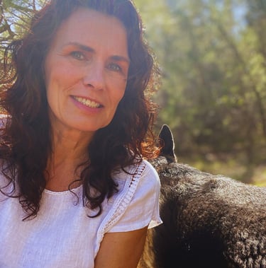 A smiling woman with curly brown hair wearing a white top outdoors with her dog.