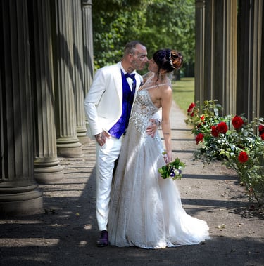 Ein Hochzeitspaar küsst sich unter einer Pergola mit Steinsäulen und einer Reihe roter Rosen; der Bräutigam trägt einen weiße