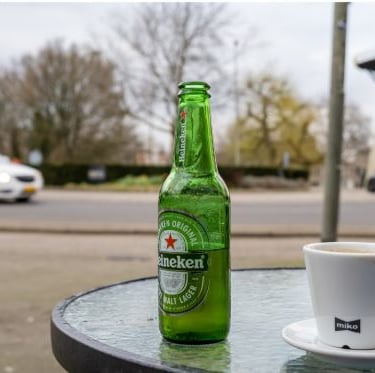 A green Heineken beer bottle and a cup of coffee on an outdoor glass table by a street.