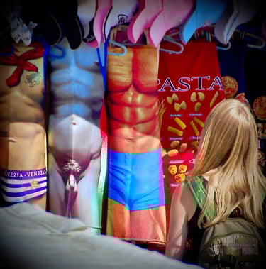 A tourist browses souvenir aprons with muscular male torso prints and pasta patterns at an outdoor Italian market stall.