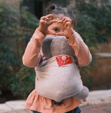 a little girl holding a stuffed animal in a babywrap carrier