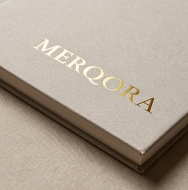 Detailed photography of a luxury brand book with gold embossed typography reading MERQORA, resting on a textured bone-colored linen surface. The lighting is moody and focused, highlighting the premium tactile quality of the materials.