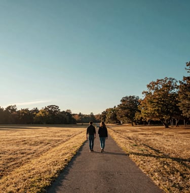 A wide-angle landscape shot of a couple walking together along a path at sunrise in a North American park, symbolizing a fresh start and shared journey. Soft morning light, palette of sandy yellow and deep teal.