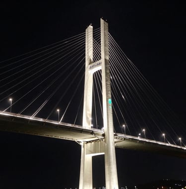 A high-contrast architectural photograph of a modern bridge structure at night, symbolizing the link between continents. The style is minimalist and premium, using deep black sky and bright off-white structural lighting with subtle sea green reflections on the metal.