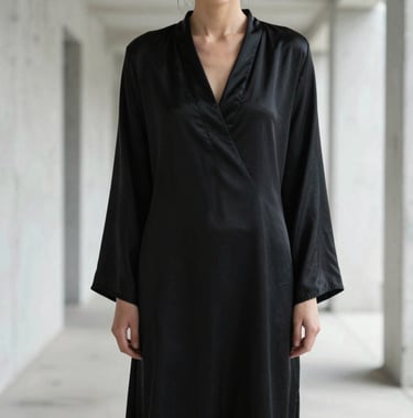 A high-fashion editorial shot of a person in a minimalist black silk garment, standing in a brightly lit white concrete corridor. Face obscured to focus on form and texture. North American / International style, soft contrast.