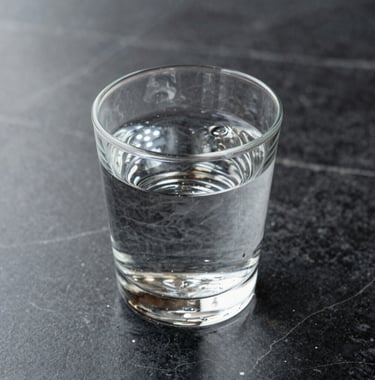 A macro shot of a single crystal glass of water on a black marble table. Sharp focus on the refractive light and the texture of the stone. Muted grey and white tones, very clean and minimalist composition.