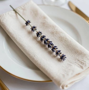 A close-up of a gold-rimmed place setting with a single sprig of dried lavender on an off-white linen napkin. North American wedding reception decor, elegant and minimalist aesthetic.