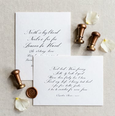 Flat lay of luxury North American / US wedding stationery on a cream linen background, featuring charcoal calligraphy, muted brown wax seals, and a few scattered cream flower petals. The composition is artistic and elegant.