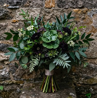 A cinematic photograph of a wedding bouquet with deep forest green foliage and dark wildflowers, set against the stone wall of an ancient castle.