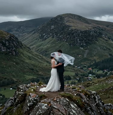 A cinematic medium shot of a wedding couple standing atop a rugged Highland peak in a Northern European / Scottish landscape. The bride's veil is caught in the wind. The lighting is moody and dramatic with deep slate and forest green tones in the background hills.