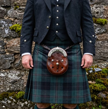 Close-up photography of a groom in a traditional kilt with a leather sporran, standing against the weathered stone walls of a Northern European / Scottish castle. Muted green moss highlights the stones, and the lighting is professional and moody.