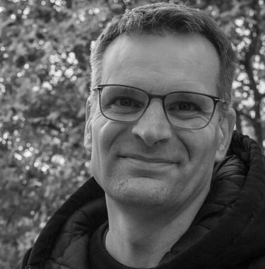 Black and white portrait of a smiling middle-aged man with glasses outdoors against bokeh trees.