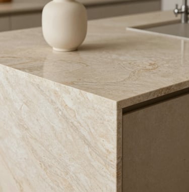 A close-up architectural photograph of a bespoke kitchen island. The surface is a light stone beige marble with delicate warm grey veining. A single minimalist soft cream vase sits on the counter. The lighting is soft and natural.