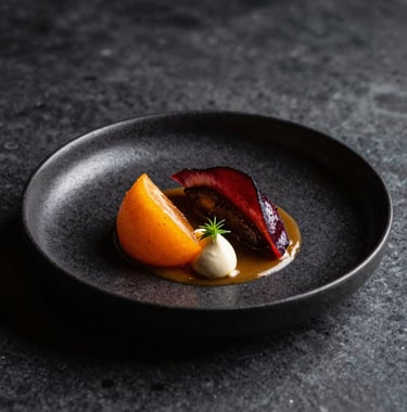 A high-end, artistic food photograph of a modern gourmet dish served in a Scandinavian-style restaurant. The lighting is dramatic, highlighting textures and colors like orange and deep reds against a black ceramic plate.