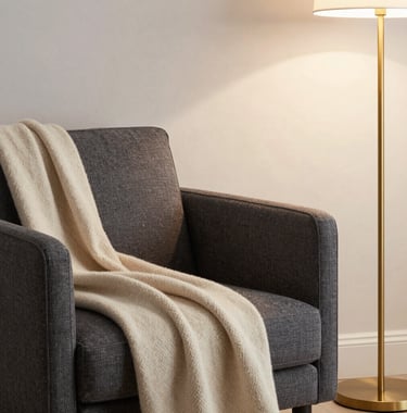 A sophisticated reading nook with a charcoal gray armchair, a warm cream-beige cashmere throw, and a soft gold floor lamp in a light-filled corner. International / High-end.