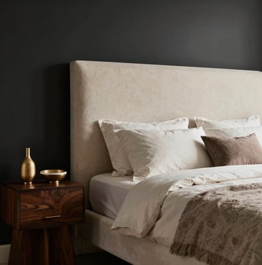 Luxury bedroom interior designed with ivory and taupe textiles. A large upholstered headboard against a matte black feature wall, soft warm lighting, and minimalist South Asian / Indian decorative elements on a dark wood side table.