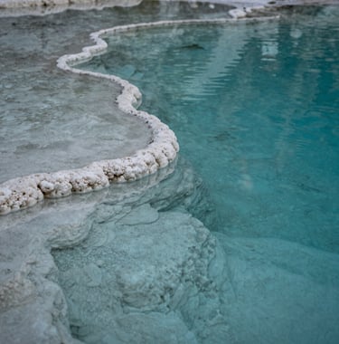 A close-up photograph of a natural mineral pool at Hierve el Agua, with crystal clear turquoise water reflecting a soft white stone edge. The lighting is soft and contemplative, highlighting the textures of the mineral deposits.