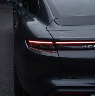 A sophisticated shot of a Porsche Taycan's rear light bar glowing in a dark environment. The car is obsidian black, and the light reflects softly off the metallic surfaces. The composition is clean and centered.
