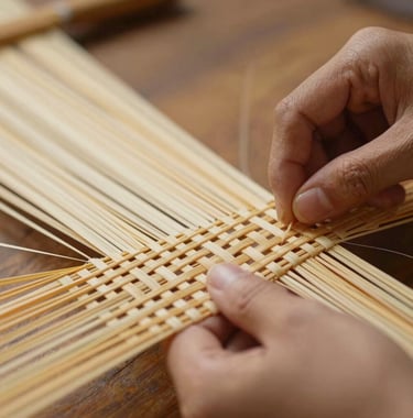 A close-up photograph of artisan hands working with warm beige bamboo fibers, weaving them into a sophisticated pattern. The lighting is soft and golden, emphasizing the craftsmanship and natural elegance of the materials.
