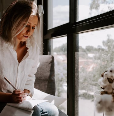 Woman in a white linen shirt writing in a notebook by a bright window.