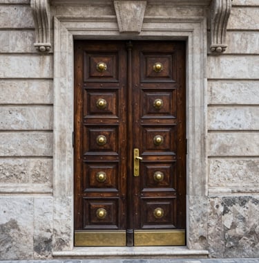 Architectural detail photography of a carved dark wooden door with brass fittings, set into a light grey stone wall of a historic Southern European building.