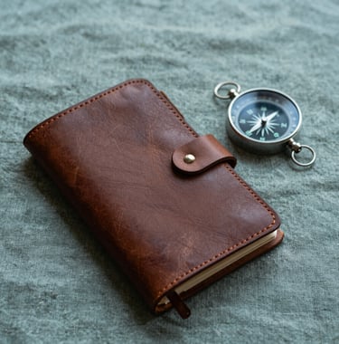 A close-up of a hand-crafted leather travel journal and a compass on a sage teal linen surface. The lighting is evocative and sophisticated, suggesting a personalized and exclusive journey in Latin America.