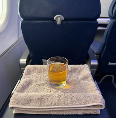 A first-person view of a luxury business class seat with a welcome drink and a warm towel. The cabin lighting is soft and modern, using midnight blue tones to convey comfort and exclusivity.