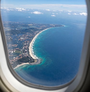 Aerial view of a South American coastline from an airplane window, showing deep blue ocean and white sandy beaches. Bright, sunny daylight, high-end travel photography style.