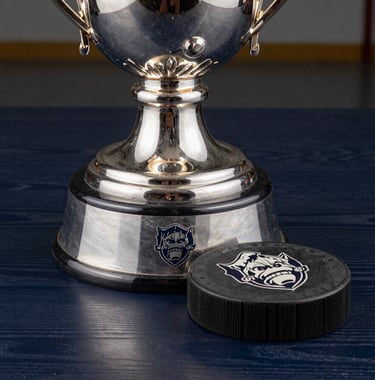 Close-up of a silver collegiate hockey trophy on a dark navy wooden table, etched logos catching the studio light, professional and prestigious feel.