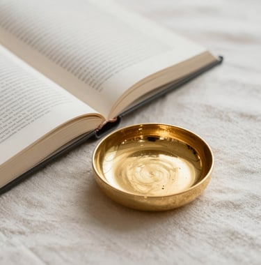 A still life photograph featuring an open book and a small gold jewelry dish on a soft off-white linen surface. The atmosphere is calm and elegant, representing focus and self-care in a South American / Brazilian aesthetic.